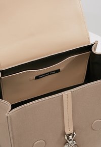 Béžová kabelka s hladkým koženým interiérem, látkovým exteriérem a stříbrnými doplňky. Obsahuje štítek s logem a otevřenou kapsu uvnitř.