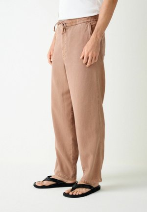 Person trägt locker sitzende hellbraune Hose mit Kordelzug in der Taille und schwarze Flip-Flops, steht vor einem schlichten hellen Hintergrund.