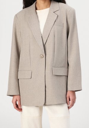 Beige single-button blazer with notched lapels, flap pockets, and a welt chest pocket worn over a white top and light pants.