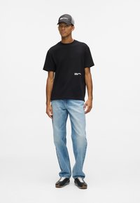 Fekete pamut póló rövid ujjakkal és fehér logóval, világoskék farmernadrággal és fekete cipővel, fehér fűzőkkel. Szürke sapka.