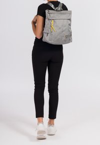 Grauer Rucksack mit strukturiertem Oberflächen, vorderer Reißverschlusstasche, gelbem Reißverschlussanhänger und zwei Seitentaschen. Am Rücken getragen mit silbernen Trägern.