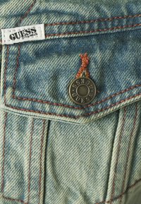 Denim tkanina svijetloplave boje i vidljive teksture. Sadrži narančaste šavove, nabiranu džepu i metalnu dugmad s ugraviranim "GUESS".