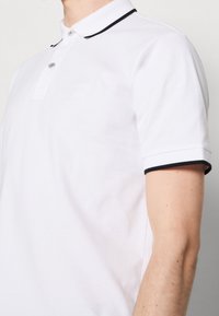 Polo blanc à manches courtes avec une bordure noire sur le col et les bords des manches, porté par une personne visible des épaules jusqu'au milieu du torse.