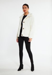 Hvid faux fur jakke med spidse kraver, guld knapper og to forlommer, parret med sorte leggings og ankelstøvler.