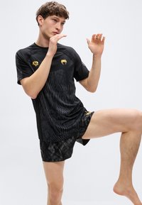 Μαύρο αθλητικό πουκάμισο με χρυσές λεπτομέρειες, που διαθέτει ανάγλυφο σχέδιο. Συνδυάζεται με μαύρο σορτς με αντίστοιχο μοτίβο.