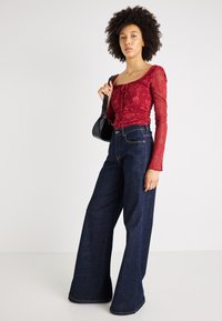 Κόκκινο δαντελένιο τοπ με μακριά μανίκια και τετράγωνη λαιμόκοψη, συνδυασμένο με ψηλόμεσα ναυτικά τζιν σε φαρδιά γραμμή με απαλό ύφασμα.