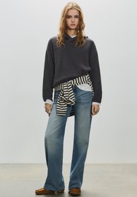 Szürke kötött pulóver, alatta fehér galléros ing, deréknál csomózott csíkos anyag, bőszárú kék farmernadrág, barna mokaszin cipő.