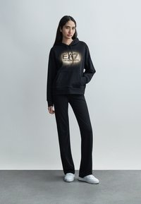 Γυναίκα που στέκεται μπροστά σε απλό τοίχο φορώντας μαύρο φούτερ Emporio Armani, μαύρο παντελόνι και λευκά αθλητικά παπούτσια, με το αριστερό της χέρι στην τσέπη.