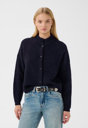 Kvinde iført navyblå knappet cardigan og lyseblå jeans med sort bælte, stående mod en helt lys baggrund.