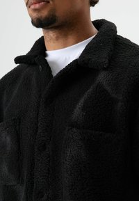 Zwarte fleecejas met een kraag, knoopsluiting en twee zakken aan de voorkant. De textuur lijkt zacht en gestructureerd.