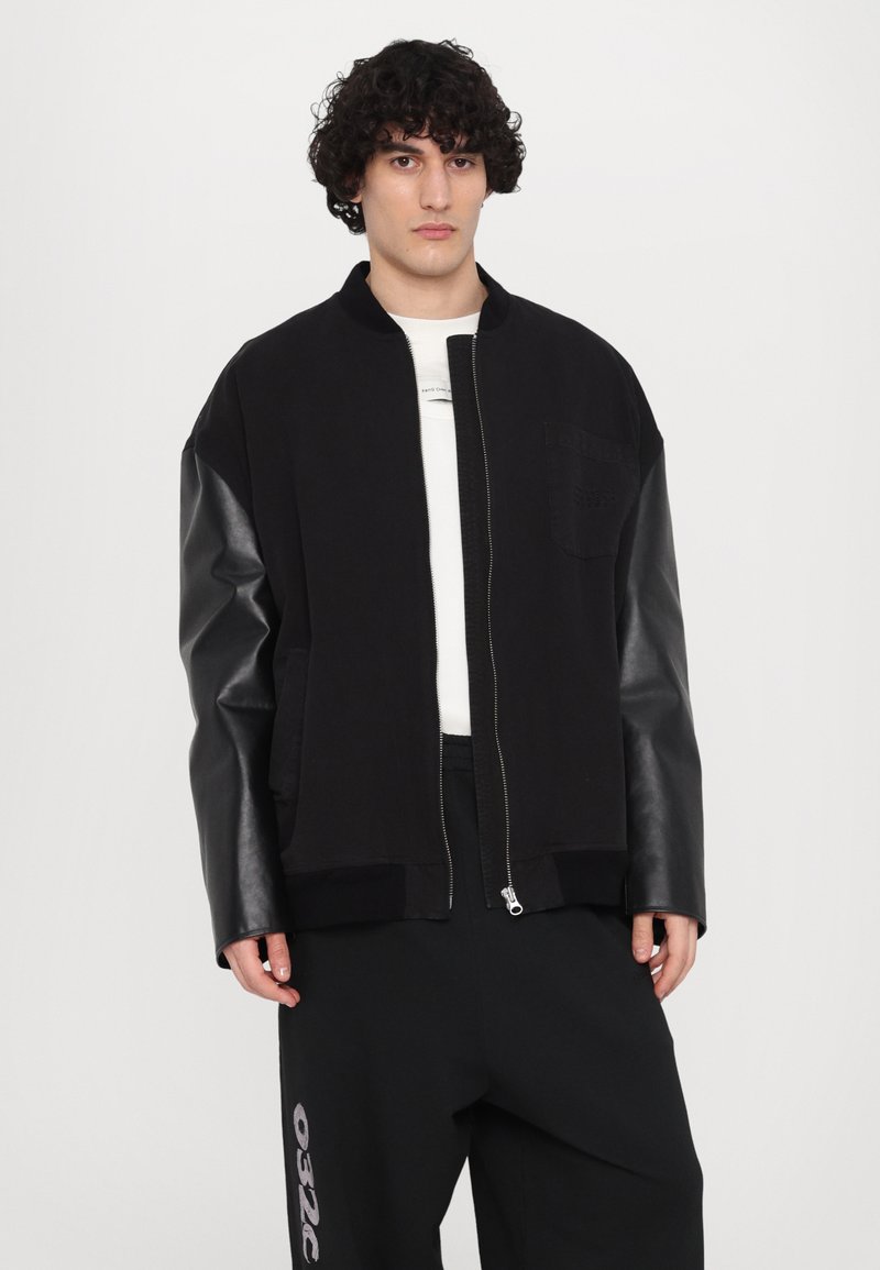 Schwarze Jacke mit kontrastierenden Lederärmeln, Reißverschluss und einer Tasche auf der Brust. Getragen über einem weißen Hemd und schwarzen Hosen.