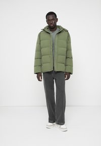 Mann in olivgrünem Steppjacke, grauem T-Shirt, dunkelgrauen weitem Jeans und weißen Sneakers, der vor einem weißen Hintergrund steht.