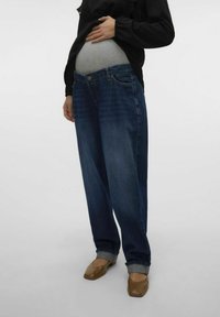 Modre denim nosečniške kavbojke z ohlapnim krojem, ki vključujejo raztegljiv pas, zavite manšete in obrabljen videz. V kombinaciji z rjavimi čevlji.