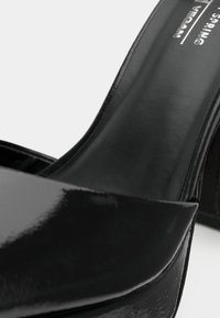 Μαύρο vegan δερμάτινο πέδιλο με ψηλό τακούνι, γυαλιστερή υφή, σχεδιασμό με φαρδιά λουριά, ανοιχτή μύτη και λεία εσωτερική επένδυση.