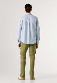 Hellblaues Langarmhemd mit Knopfmanschetten, kombiniert mit olivgrünen Hosen. Die Rückansicht zeigt einen abgerundeten Saum und einen lockeren Schnitt.