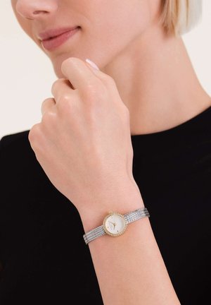 Montre-bracelet en mailles métalliques argentées avec un cadran blanc, des accents dorés et une lunette décorative sertie de pierres, portée sur une main.