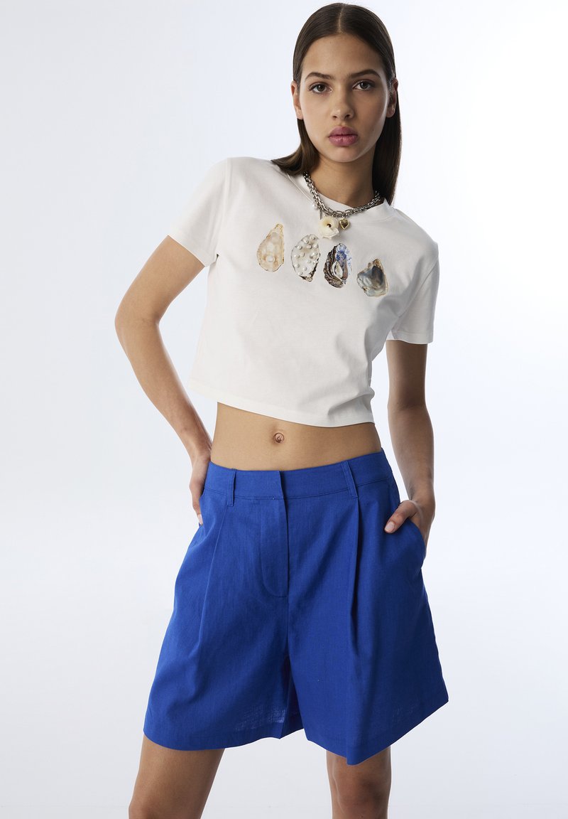 T-shirt bianco corto con una grafica di cinque conchiglie. Pantaloni corti in lino blu con vestibilità ampia e pieghe frontali.