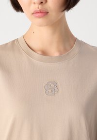 Primo piano di una persona che indossa una maglietta beige a girocollo con un logo doppio "B" impreziosito da strass al centro del petto.