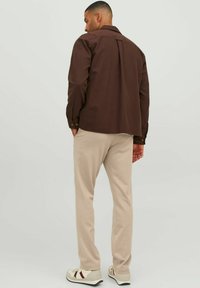 Camisa marrón de botones con cuello, corte relajado y pliegue en la parte de atrás, combinada con pantalones color crema y zapatillas blancas con acentos rojos.