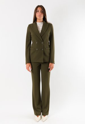 Donna che indossa un completo doppiopetto verde oliva con ampi revers, top in pizzo bianco e scarpe bianche con punta affusolata, in piedi davanti a uno sfondo uniforme.