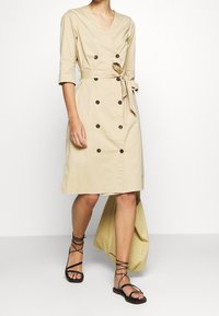 Beige Wickelkleid mit V-Ausschnitt, dreiviertel langen Ärmeln, doppelt knöpfbaren Verschlüssen, einem selbstbindenden Gürtel und einem knielangen Saum. Schmale schwarze Sandalen.