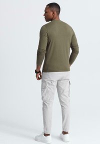 Man som står med ryggen mot kameran, klädd i en olivfärgad långärmad skjorta, ljusgrå cargo-byxor, vita sneakers och en svart armbandsklocka, mot en vit bakgrund.