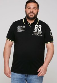 Black polo shirt with yellow accents, featuring printed text and numbers. Short sleeves with contrasting collar. Made from cotton blend fabric.