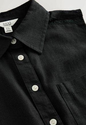 Camicia di lino nera con colletto classico, caratterizzata da bottoni bianchi, una tasca sul petto e un delicato inserto di pizzo sulla spalla.