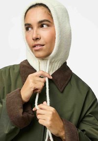 Capuche en tricot crème, texture côtelée, avec deux cordons blancs. Associée à une veste surdimensionnée verte dotée d'un col en velours côtelé marron.