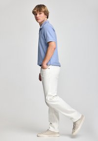 Ljusblå polo med klassisk krage, ihop med vita jeans och beige sneakers, stående i profil mot en neutral bakgrund.