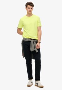 Camiseta amarilla brillante de manga corta, jeans oscuros, suéter gris atado a la cintura, zapatillas blancas con acentos negros, con un estilo casual.