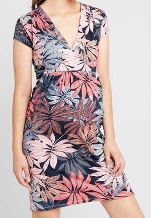 Frau trägt ein kurzärmeliges V-Ausschnitt-Kleid mit einem tropischen Blattmuster in Pink, Rot und Blau, posiert mit einer Hand auf der Hüfte.