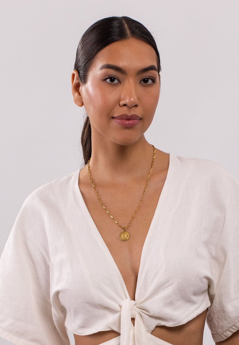 Goldene Kettenhalskette mit einem runden Anhänger, getragen über einem weißen, vorne gebundenen Oberteil. Glatte Textur, geschichtetes Design und mittlere Dicke.