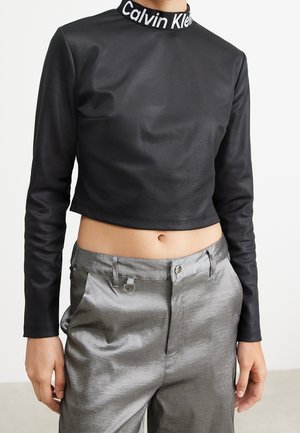 Person trägt ein schwarzes langärmliges, bauchfreies Oberteil mit dem Schriftzug "Calvin Klein" am Kragen und glänzende silberne High-Waist-Hosen.