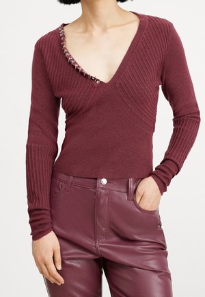 Femme portant un pull en maille côtelée à col en V profond avec des détails œillets argentés sur un côté et un pantalon en cuir taille haute assorti couleur bordeaux.
