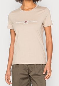 Žena v béžovém bavlněném tričku s krátkým rukávem, na kterém je logo a nápis Tommy Hilfiger, v kombinaci se zelenými kalhotami, stojící na bílém pozadí.