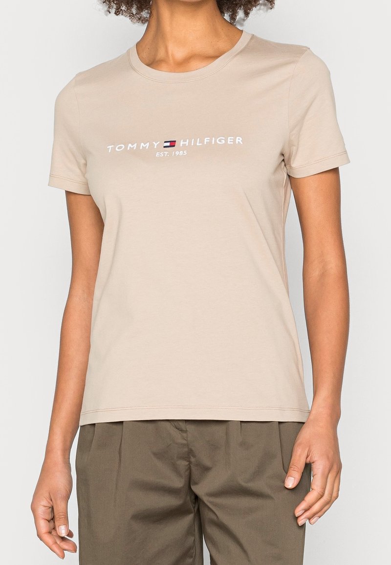 Žena v béžovém bavlněném tričku s krátkým rukávem, na kterém je logo a nápis Tommy Hilfiger, v kombinaci se zelenými kalhotami, stojící na bílém pozadí.