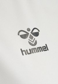 Czarne stylizowane logo pszczoły nad słowem „hummel” drukowane na białej tkaninie, pokazujące znak marki na materiale odzieżowym.