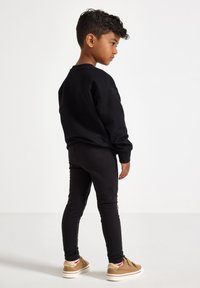 Sort sweatshirt, sorte leggings og brune sko. Billedet er taget fra siden, hvilket fremhæver et afslappet look med en glat tekstur og en uformel stil.