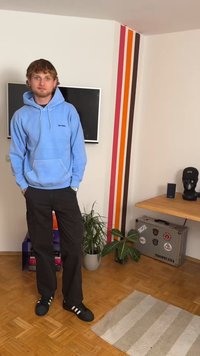 Lyseblå hettejakke med frontlomme og logo, svarte cargobukser og svarte joggesko. Innendørs setting med planter og fargerike veggdetaljer.