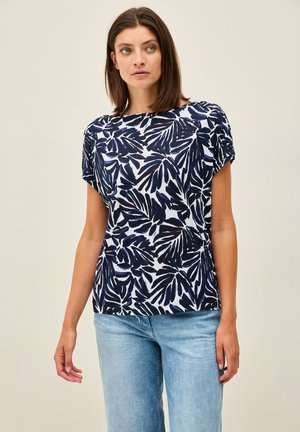 Navy and white short-sleeve top featuring a leaf pattern, round neckline, and relaxed fit, paired with light blue jeans.