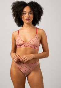 Kvinna med lockigt hår som bär genomskinlig nude-lingerie med röda hjärtan, stående mot en enkel ljusgrå bakgrund.