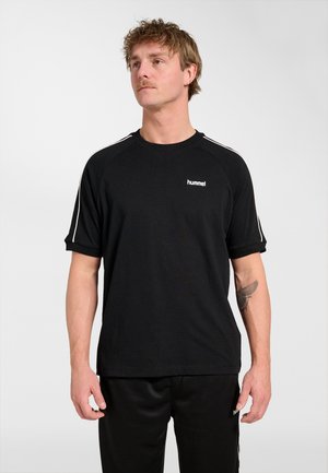 Mand med overskæg iført en sort hummel t-shirt med hvide striber på ærmerne og matchende sorte bukser, stående mod en ensfarvet baggrund.