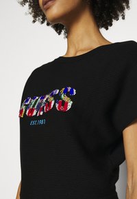 Schwarzer kurzärmeliger Rippstrickpullover mit mehrfarbigem Pailletten-Text und Akzenten in Rot, Blau und Grün, mit "EST.1981" in Blau.