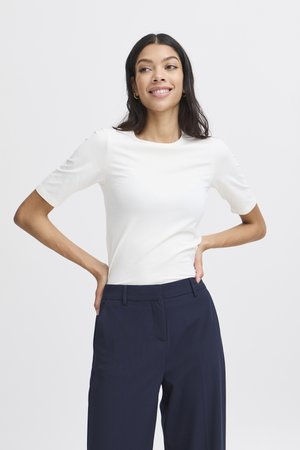Femme aux longs cheveux foncés, portant un haut blanc ajusté à manches courtes et un pantalon bleu foncé, debout les mains sur les hanches et souriante.