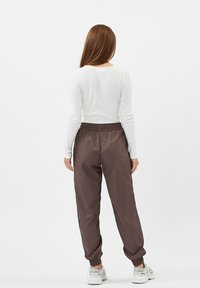 Brune, lette joggers med elastisk talje og manchetter, parret med en tætsiddende hvid bluse med lange ærmer, set fra bagsiden.