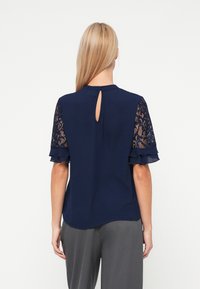 Námornícky modrá blúzka s čipkovými krátkymi rukávmi, volánovými okrajmi a detailom v tvare kľúčovej dierky na zadnej strane. Mäkká tkanina a uvoľnený strih. V kombinácii s šedými nohavicami.