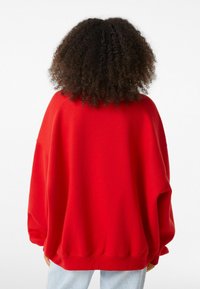 Sweat-shirt rouge surdimensionné avec épaules tombantes, poignets côtelés et texture douce. Le dos présente un design lisse et une coupe décontractée.