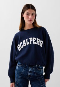 Marineblå sweatshirt med "SCALPERS" i store hvide bogstaver. Har en rund halsudskæring, sænkede skuldre og en afslappet pasform. Bæres med blå jeans.