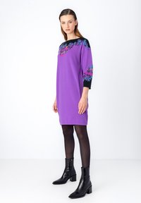 Violetti mekko mustilla olkaimilla, jossa on kukkaniittikuvioita. Malli käyttää mustia nilkkureita ja sukkahousuja, seisten valkoisella taustalla.